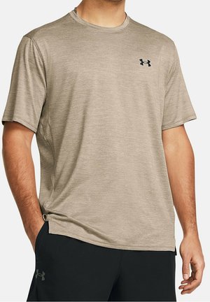 Uomo che indossa una maglietta sportiva beige a maniche corte con un logo nero Under Armour sul petto e pantaloncini neri con logo Under Armour.