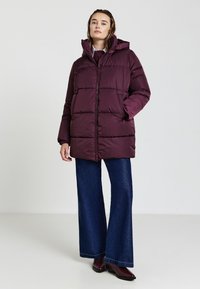 Donna che indossa un giubbotto puffo bordeaux, jeans a gamba larga blu scuro e stivali bordeaux, in piedi su uno sfondo bianco semplice.