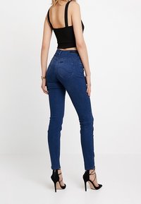 Vaqueros pitillo azul de talle alto en denim elástico, con diseño de bolsillos traseros y acabado liso. Combinados con tacones negros de tiras.