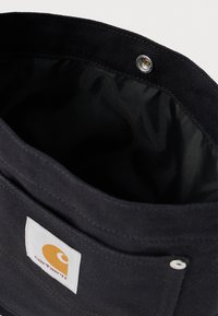 Borsa in tessuto Carhartt nera con patch rettangolare bianco, chiusura a pressione e fodera interna testurizzata visibile.