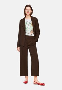 Brauner maßgeschneiderter Anzug mit weit geschnittenen Hosen und passendem Blazer. Ein grafisches weißes T-Shirt mit floralen Designs. Rote flache Schuhe mit Schleifen.