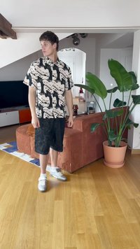 Kortärmad skjorta med svart och beige abstrakt mönster, kombinerad med mörka shorts. Vita sneakers, inomhusmiljö med växt och möbler.