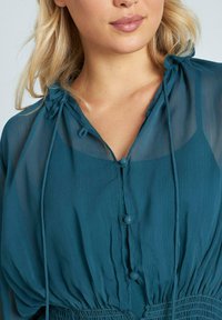 Blusa trasparente color teal con scollatura regolabile tramite coulisse, chiusura con bottoni e vita elastica, caratterizzata da una superficie testurizzata e delicate pieghe.