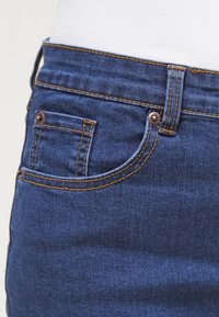 Indigo džíny z denimu s přední kapsou, měděnými detaily a kontrastním zlatým prošíváním. Navrženy v úzkém střihu s hladkou texturou.