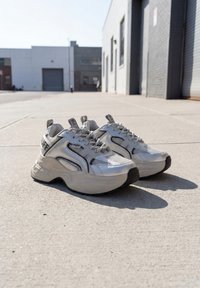 Zilveren en witte chunky sneakers met zwarte zolen geplaatst op een betonnen stoep buiten industriële gebouwen op een zonnige dag.
