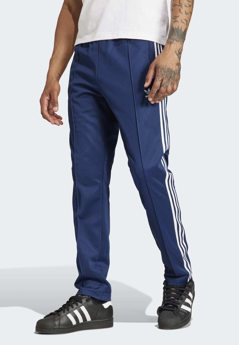 Mørkeblå sportsbukser med tre hvide striber ned ad hver side, lavet af glat stof; har en elastisk talje og et lille logo.