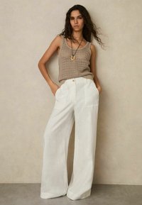 Beiges, gestricktes Tanktop kombiniert mit weißen Hosen mit weitem Bein. Die Hose hat einen Knopfverschluss und Seitentaschen. Glatte Stoffstruktur.