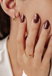 Orecchino a cerchio dorato e anello dorato intrecciato. L'orecchino ha una forma rotonda; l'anello presenta un design liscio e intrecciato. Le unghie sono dipinte di un viola scuro.