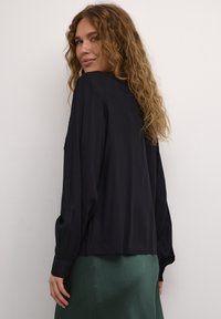 Blouse noire à manches longues avec une coupe ample, dotée de poignets boutonnés et d'une texture lisse, associée à une jupe verte texturée.