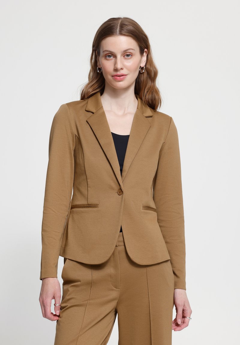 Femme aux cheveux bruns ondulés portant un blazer ajusté beige et un pantalon assorti sur un haut noir, debout devant un fond clair uni.
