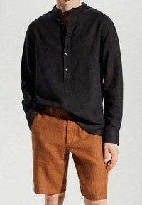 Persona vistiendo una camisa negra de manga larga abotonada metida dentro de unos pantalones cortos de color naranja quemado hasta la rodilla, de pie contra un fondo simple.