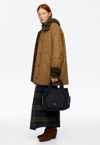 Młoda kobieta o rudych włosach nosi brązową kurtkę z ko�żuszkiem, zielony sweter, kraciastą spódnicę, brązowe zamszowe kozaki i trzyma czarną torebkę na suwak.