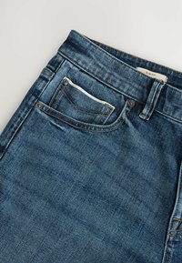 Úzke modré džínsy vyrobené z denimu. Majú zapínanie na gombík a zips, dve predné vrecká s detailným prešívaním a štítok na opasku.