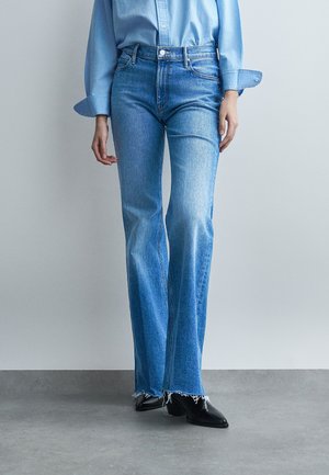 Személy világoskék farmert visel, nyers szegélyekkel, belebújtatott kék inggel és fekete hegyes orrú bokacsizmával, szürke padlón állva.