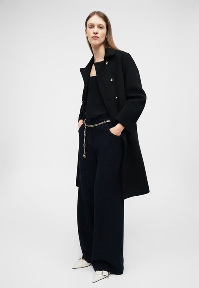 Frau steht und trägt einen schwarzen Mantel, schwarze weite Hose mit Kettengürtel, schwarzes Oberteil und weiße spitze Schuhe vor einfachem Hintergrund.