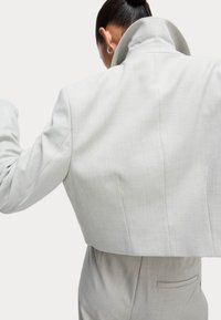 Beskuren ljusgrå blazer med hög krage, strukturerade axlar och synliga sömdetaljer, parat med en slät tygtextur.
