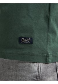 Tmavě zelené bavlněné tričko s malou námořnicky modrou štítkem s nápisem "PWrnd 73". Látka má hladkou texturu a je olemovaná na spodní straně.