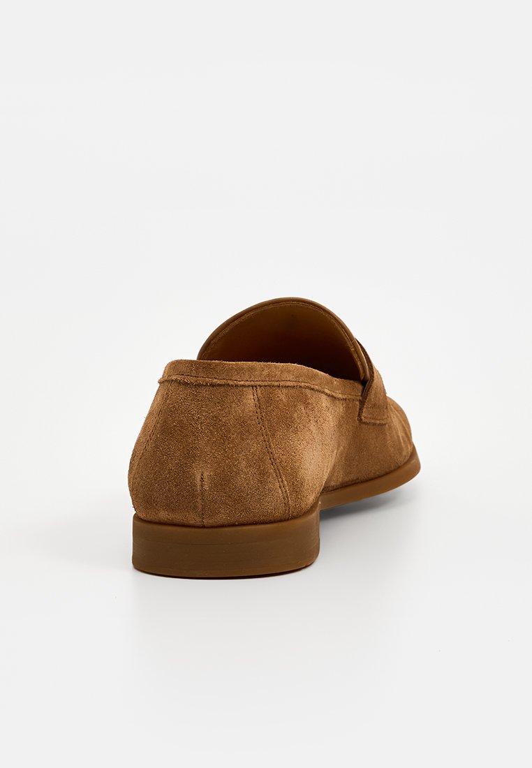 Mocasin de ante marrón con tacón bajo, visto desde la parte trasera sobre un fondo blanco.