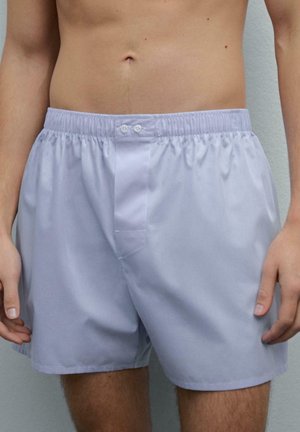Bare overkrop af en person iført lysegrå boxershorts med elastisk talje og to knapper på frontlynlåsen mod en ensfarvet baggrund.