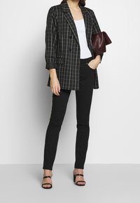 Blazer a cuadros en blanco y negro, diseño de doble botonadura, combinado con una camiseta sin mangas blanca, jeans skinny negros y sandalias de tacón negras.