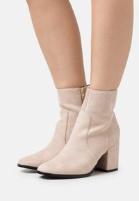 Bottes beiges en daim montant à la cheville, avec un bout pointu, une surface texturée et un talon bloc de hauteur moyenne. Détail zippé à l'arrière.