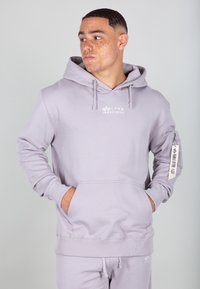 Svetlo šedá mikina s kapucňou vyrobená z jemného materiálu, s prednou kapsou, šnúrkami na stiahnutie a bočnou zipsovou kapsou s nášivkou. Logo na hrudi.