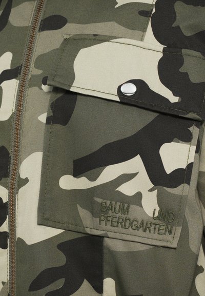 Kamo rašto audinys žalios ir smėlio spalvos atspalvių su krūtinės kišene, kuri turi sidabrinį spaudimą ir išgraviruotą tekstą "BAUM UND PFERDGARTEN."