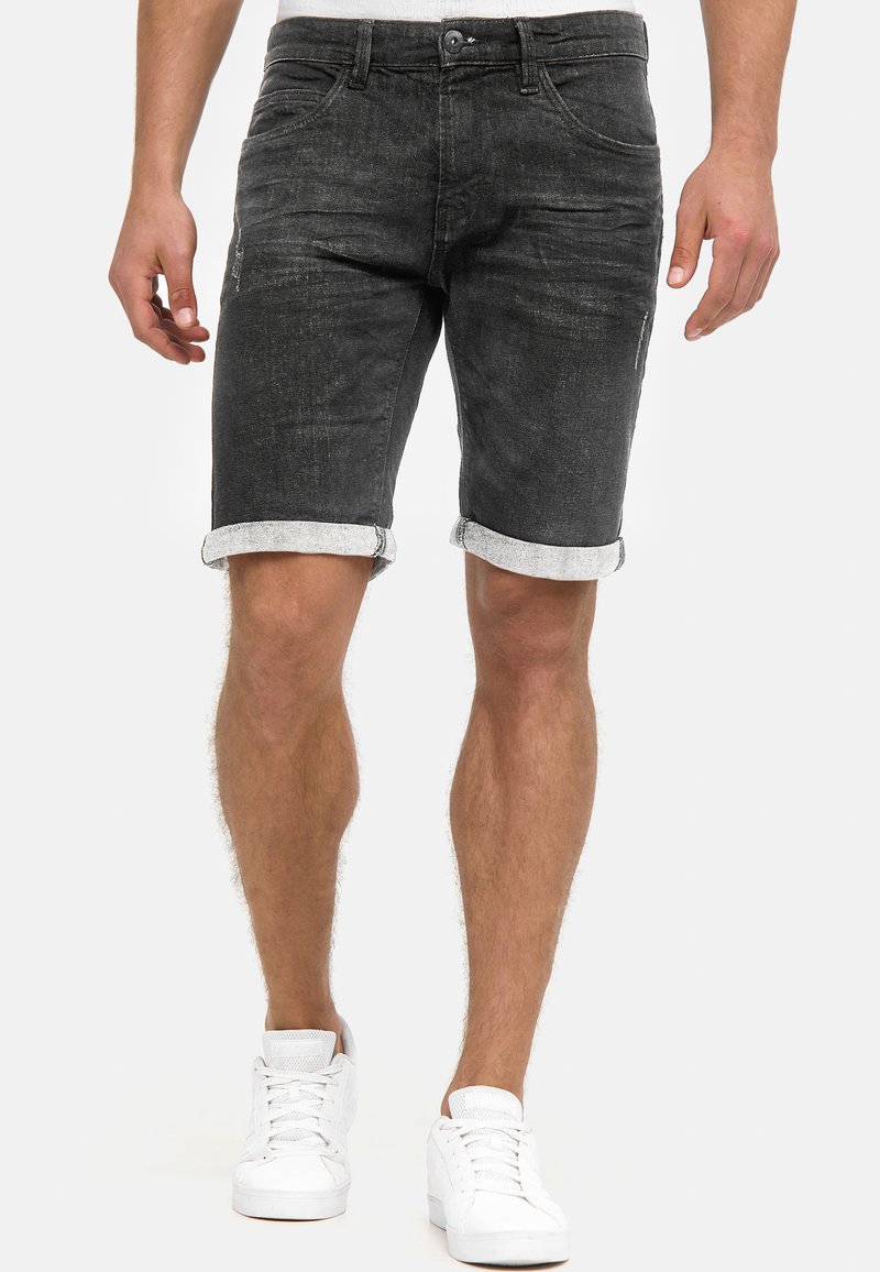 Shorts en denim noir avec revers roulés, présentant une texture délavée et des poches latérales. Associés à des baskets blanches, l'accent est mis sur une tenue décontractée.