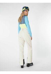 Skikleding met een lichtblauw, grijs en neongele top, crèmekleurige skibroek en zwarte skischoenen met ritsen op de benen.