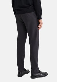 Muž v tmavě šedých kalhotách, černém svetru a černých kožených mokasínách stojí před světlým pozadím.