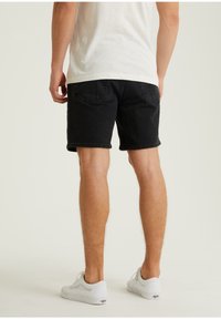 Mustat farkkushortsit suoralinjaisella leikkuulla, takataskuilla ja repaleisella helmalla, yhdistettynä valkoisiin tennareihin vaalealla taustalla.