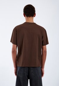 Brown cotton t-shirt with a crew neck and short sleeves, featuring a relaxed fit and smooth texture. Worn with dark denim pants.