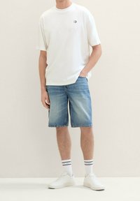 Mies, joka pukeutuu valkoiseen lyhythihaiseen t-paitaan, sinisiin denim-pitkiin shortseihin, valkoisiin raidallisiin sukkiin ja valkoisiin lenkkikengiin, seisoo yksinkertaista taustaa vasten.