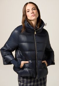 Marineblå puffjakke lavet af glat materiale, med høj krave, frontlynlås og sidelommer. Ribstrikkede manchetter fuldender designet.