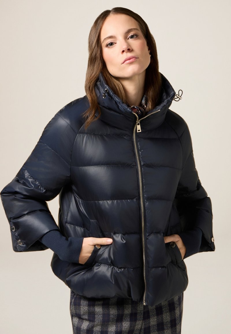 Marineblå puffjakke lavet af glat materiale, med høj krave, frontlynlås og sidelommer. Ribstrikkede manchetter fuldender designet.