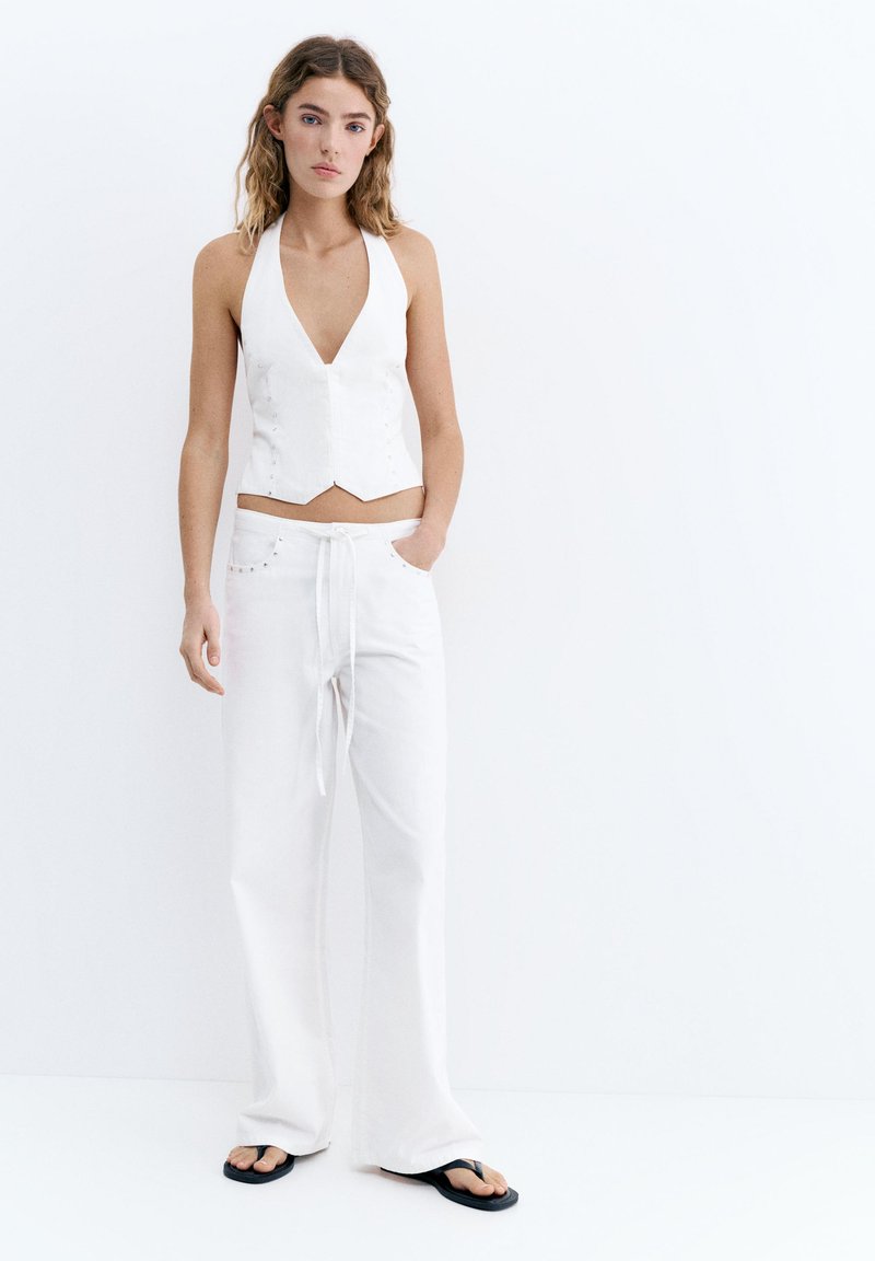 Jeune femme debout contre un mur blanc portant un gilet blanc sans manches, un pantalon blanc ample avec cordon de serrage et des tongs noires. Mains dans les poches.