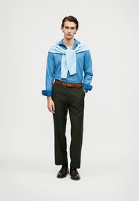 Camicia di jeans con bottoni, maglione azzurro chiaro annodato al collo, pantaloni verde scuro, cintura marrone, scarpe marroni, in piedi davanti a uno sfondo bianco.