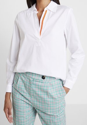 Vrouw draagt een witte blouse met lange mouwen met een oranje bies langs de open kraag en lichtblauwe geruite broek met een zwarte knoop.