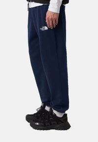 Pantalones de chándal azul marino con un logo blanco en la pierna izquierda, combinados con zapatillas deportivas negras con detalles de malla y goma.