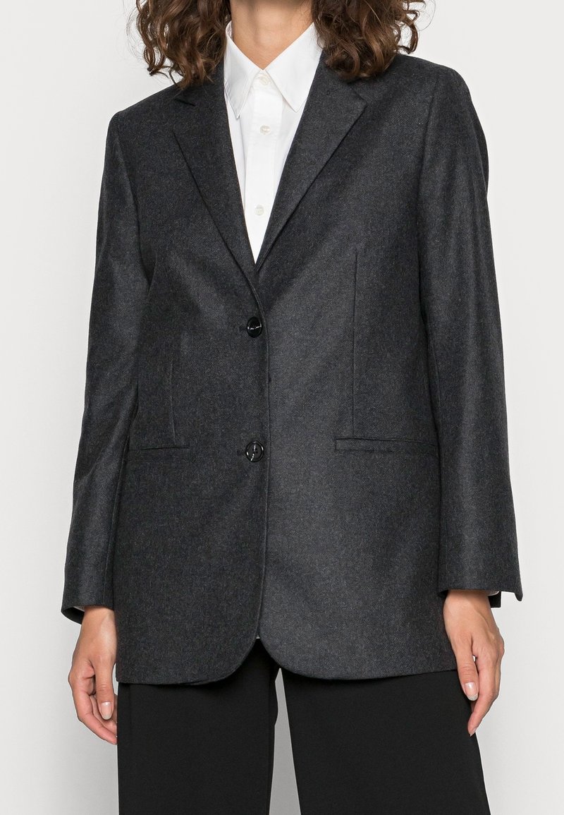 Personne portant un blazer boutonné gris foncé sur une chemise blanche à col et un pantalon noir, debout devant un fond uni.