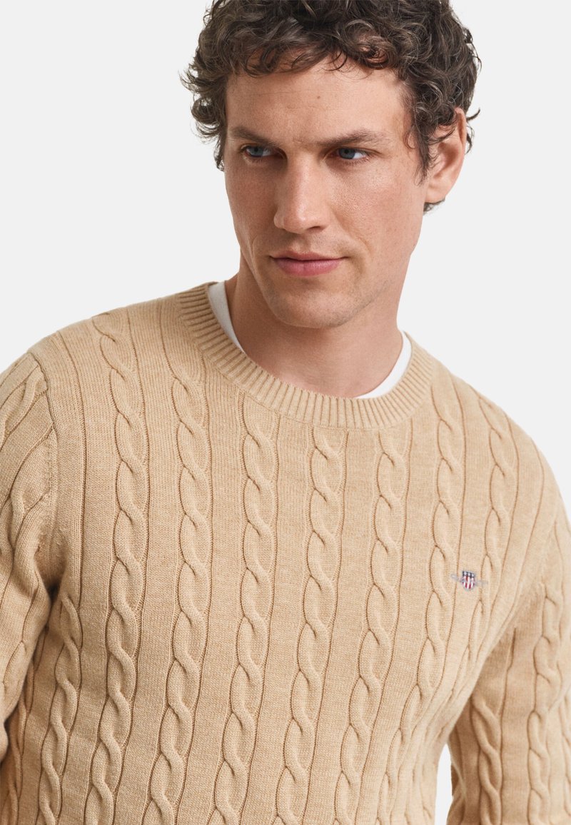 Beige Kabelstrickpullover mit rundem Halsausschnitt und gerippten Bündchen. Besitzt ein kleines Logo auf der Brust. Weicher Stoff, klassischer Schnitt.