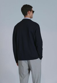 Cardigan bleu marine avec une coupe décontractée, fabriqué en tissu doux. Il possède des manches longues, un col classique, et est assorti à un pantalon gris clair.