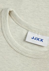 Ribbete, lys beige strikket stoff med rund hals og en blå etikett som viser "JJXX" brodert på innsiden.
