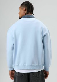 Sweat-shirt bleu clair avec un col en denim, coupe oversize, poignets et ourlet côtelés. Texture douce et design décontracté, porté sur une chemise blanche.