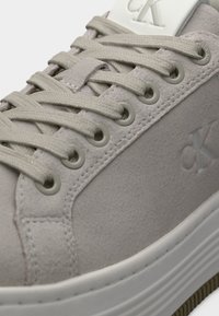 Γκρι σουέντ αθλητικό παπούτσι με επίπεδα μπεζ κορδόνια, λογότυπο στη λευκή γλώσσα και υφασμένο πέλμα με ορατό σχέδιο πέλματος. Κυκλικές οπές για κορδόνια.