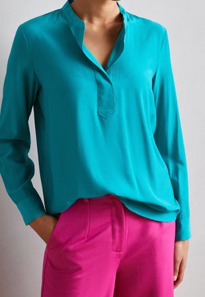 Türkise Bluse mit langen Ärmeln, Notch-Kragen und lockerer Passform, kombiniert mit rosa Stoffhosen. Sanfte Textur, dezenter Glanz und klare Linien.