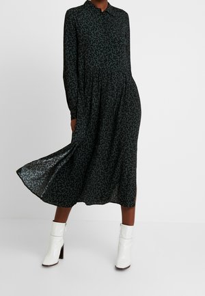 Mujer con vestido midi de manga larga y estampado oscuro, combinado con botas blancas hasta el tobillo, de pie con un lado del vestido ligeramente levantado.