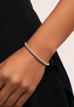 Pulsera de plata retorcida, de forma redonda, con textura suave, llevada en una muñeca sobre un fondo de prenda oscura.