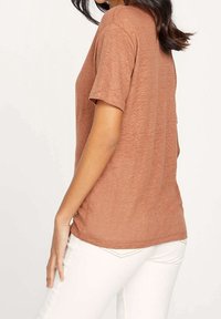 Žena ve tričku s krátkým rukávem v měděné barvě a bílých kalhotách, stojící bokem s jednou rukou na boku.