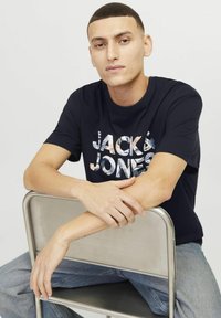 Tamsiai mėlynos medvilnės marškiniai su daugiakomponenčiu logotipo Spaudiniu "JACK & JONES." Dėvimi su šviesiai mėlynomis džinsomis, modelis sėdi ant metalinės kėdės.